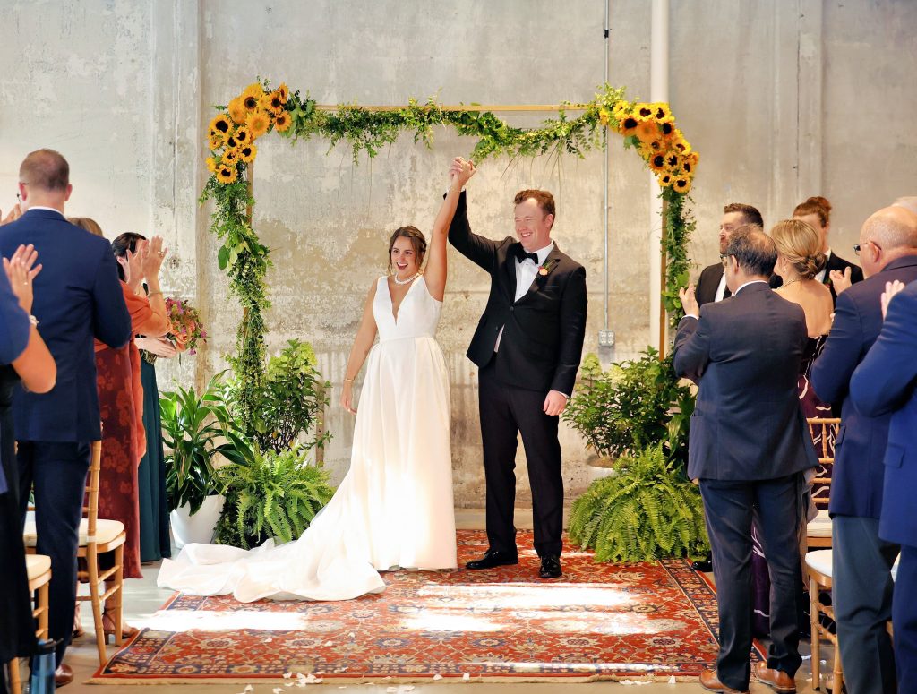A bride and groom raise their held hands against a greenery arch that features clusters of sunflowers in front of a clapping crowd.
