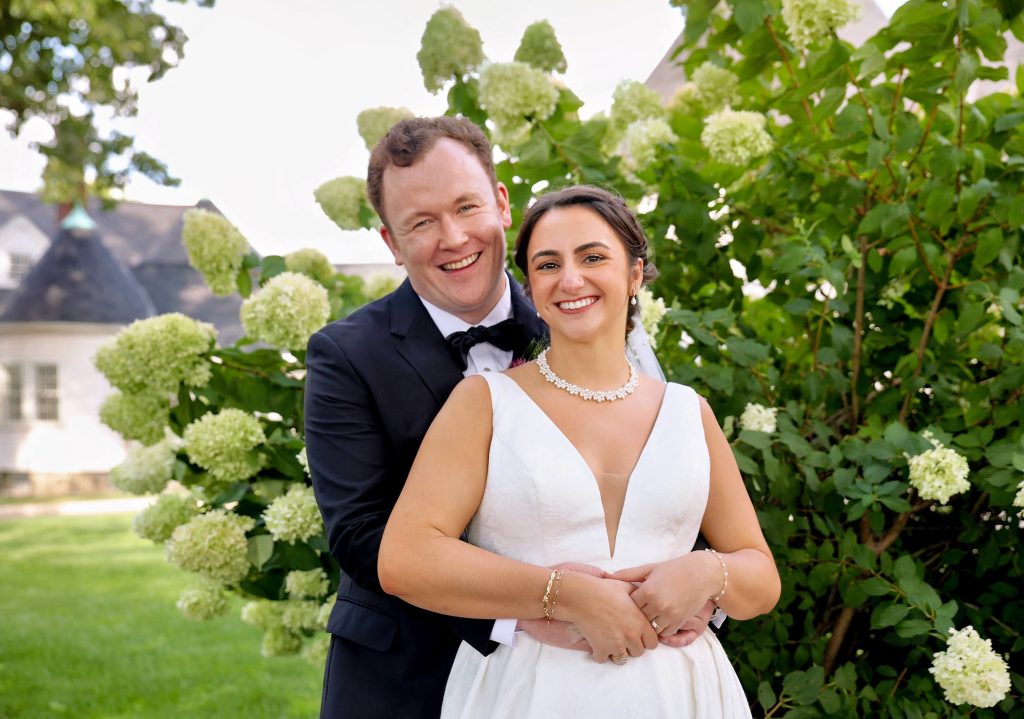 A groom holds his bride by her waist as they look at the camera, smiling, in front of a large hydrangea bush.