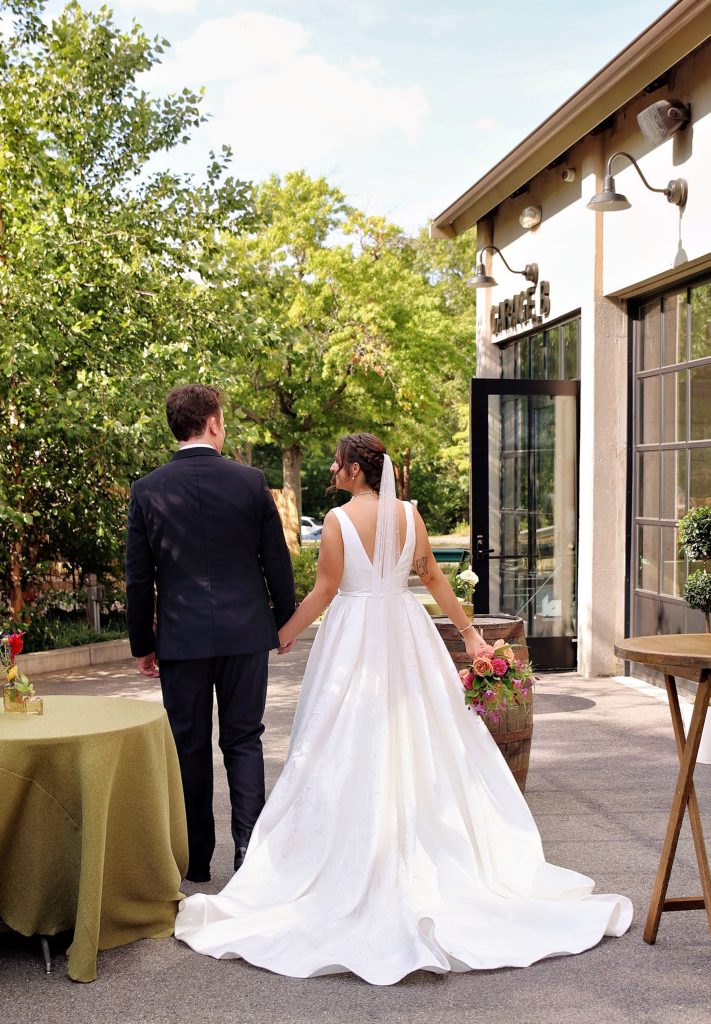 A groom and bride walk hand-in-hand away from the camera on an outdoor patio adjacent to Garage B that is arranged with barrel high-tops and low round banquet tables.