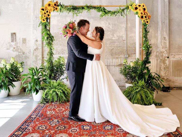 A groom and bride pose, smiling, with their arms around each other on top of a boho rug in front of a greenery arch dotted with bunches of sunflowers set against a distressed stone wall.