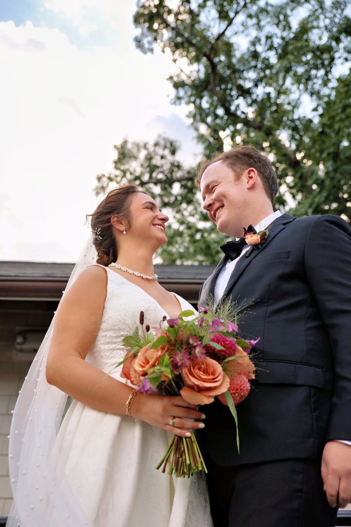A bride, holding her bouquet, and groom look at each other, smiling.