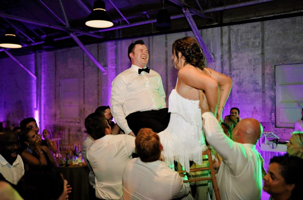 A groom and bride are raised on chairs by a group of men while other dancing wedding guests watch, smiling and laughing, in a warehouse-style venue featuring distressed stone walls, steel rafters, and pendant lighting.