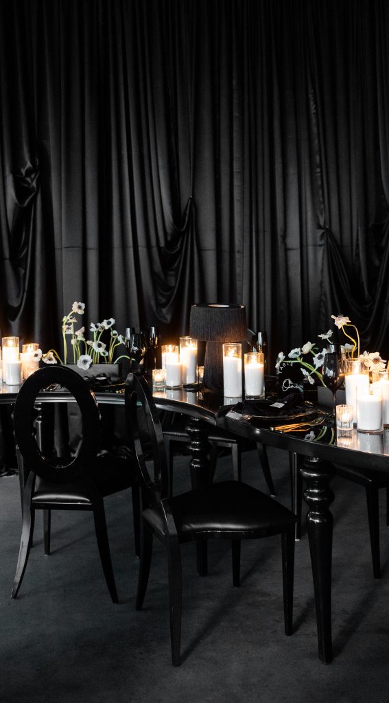 A monochromatic tablescape atop a serpentine dining table, set with battery-operated candles, fringed lamps, and arrangements of anemones against a backdrop of drapery.