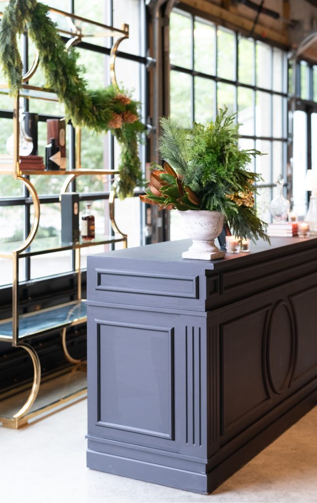 A bar, topped with an arrangement of firs, in front of a metal bar shelf set against a pair of floor-to-ceiling glass windows.