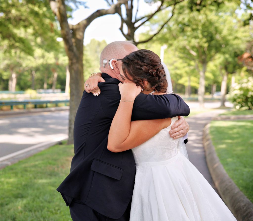 An older man and a bride hug tightly on a roadside lined with trees and grass.