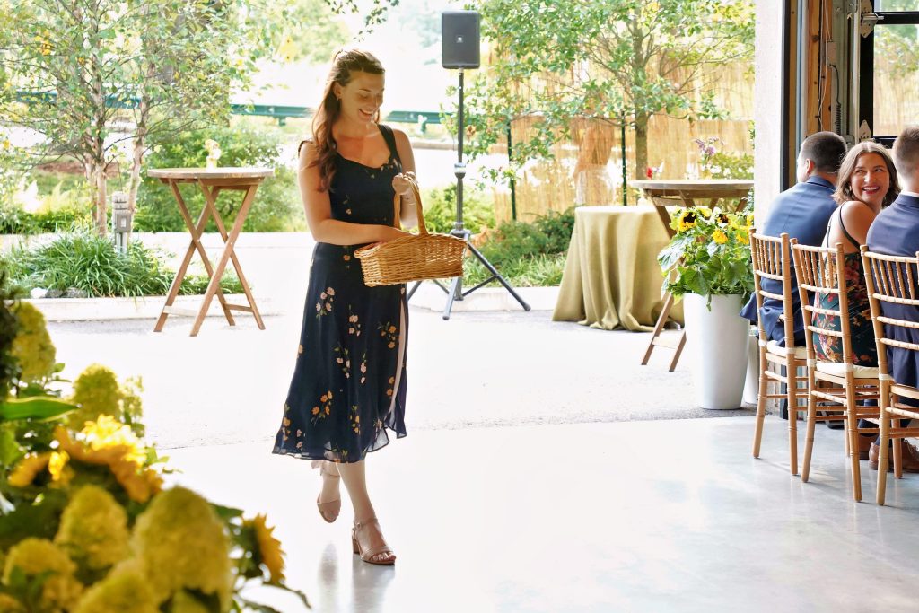 A smiling person, carrying a basket, walks from an outdoor patio into a building with polished concrete flooring as part of a wedding procession.
