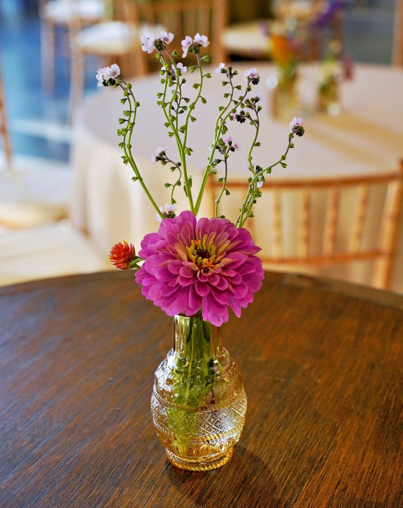 A glass bud vase of a few sparse meadow flowers sits atop a wooden surface with banquet tables and wooden Chiavari chairs in the background.