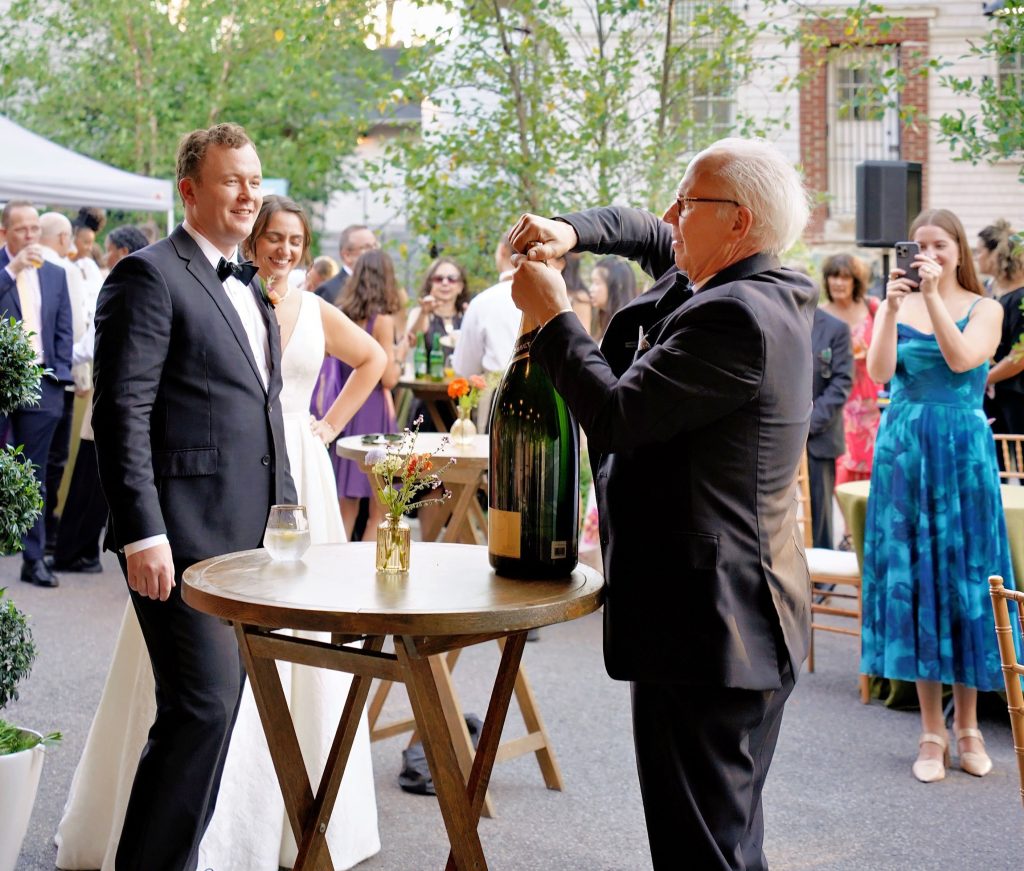 A man opening an oversized bottle of champagne in front of a smiling groom and bride as well as other wedding guests on an outdoor patio.