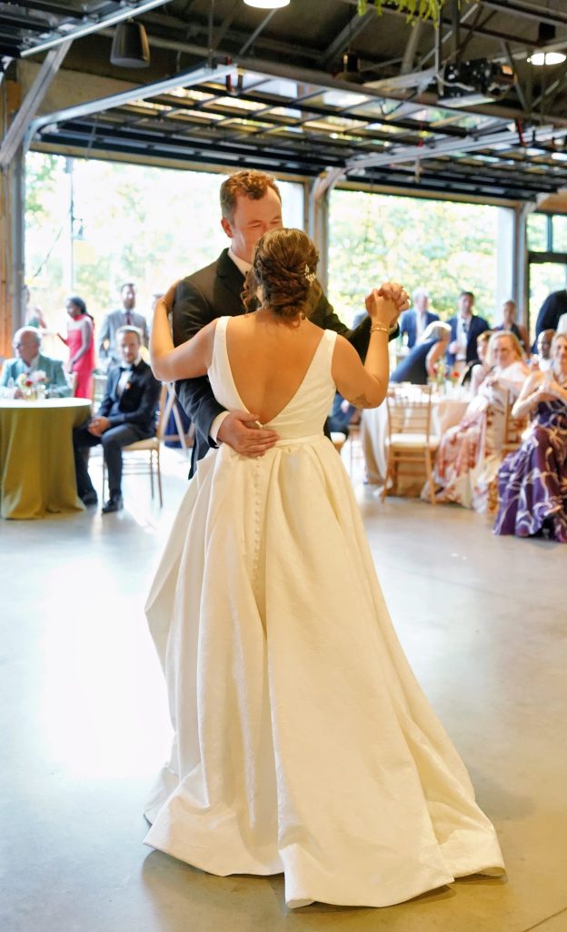 A groom and bride dance upon a polished concrete floor for wedding guests seated and standing around round banquet tables against a backdrop of opened glass overhead doors that provide a view to the outside trees.
