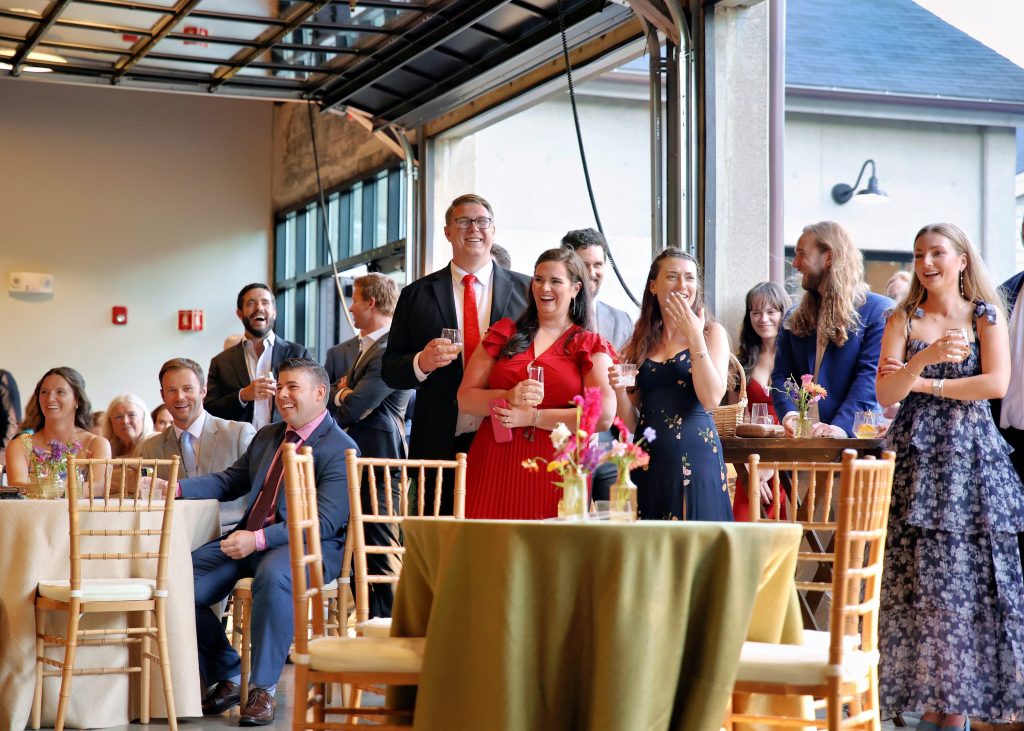 Smiling and laughing wedding guests sitting and standing around round banquet tables dressed with canvas linens and surrounded by wooden Chiavari chairs underneath opened overhead glass doors.