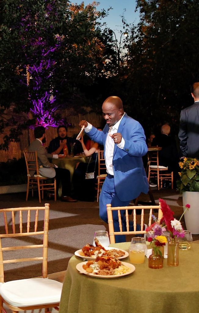 At dusk, a man in a suit dances on a patio arranged with low round banquet tables dressed with canvas linens and wooden Chiavari chairs.
