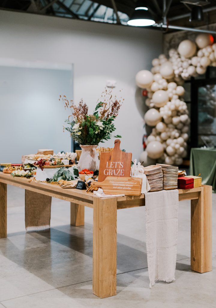 A wooden grazing table -- complete with various charcuterie items and a dried floral arrangement -- in a modern-industrial venue decorated with a balloon arch.