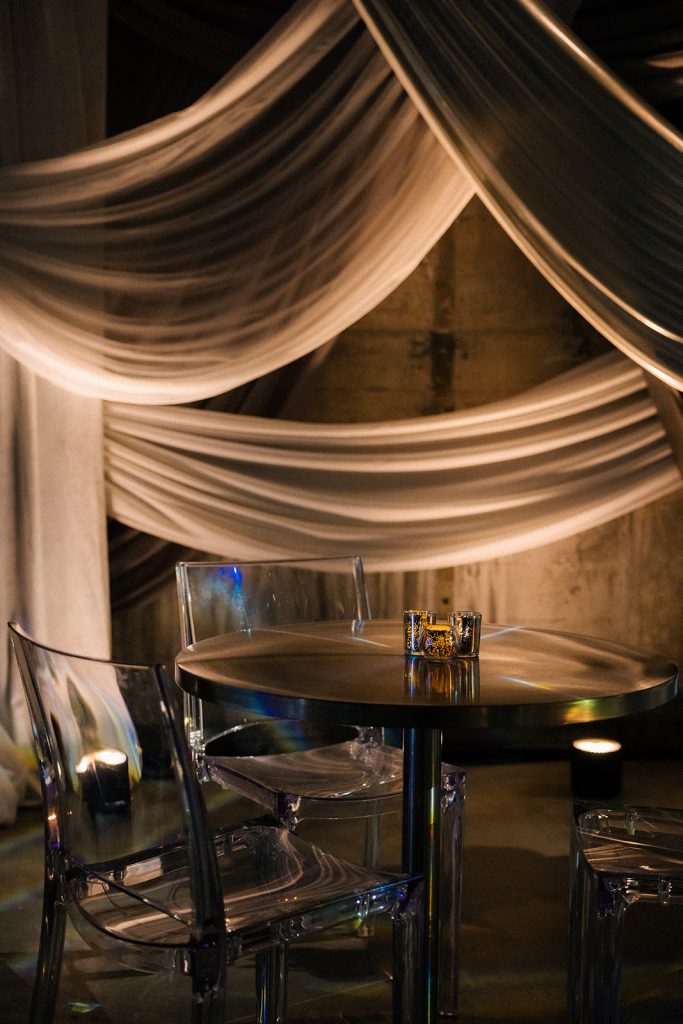 Lucite chairs at a cocktail table in front of a draping arrangement set against a distressed stone wall in warm, dim lighting.
