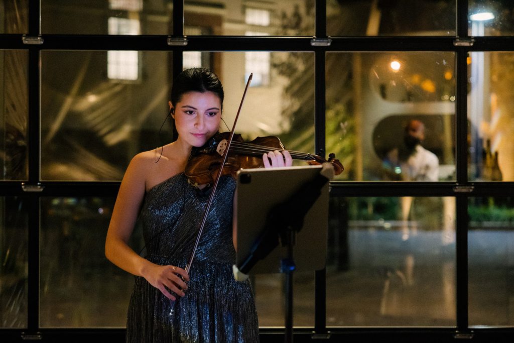 An electric violinist plays against a large glass window.
