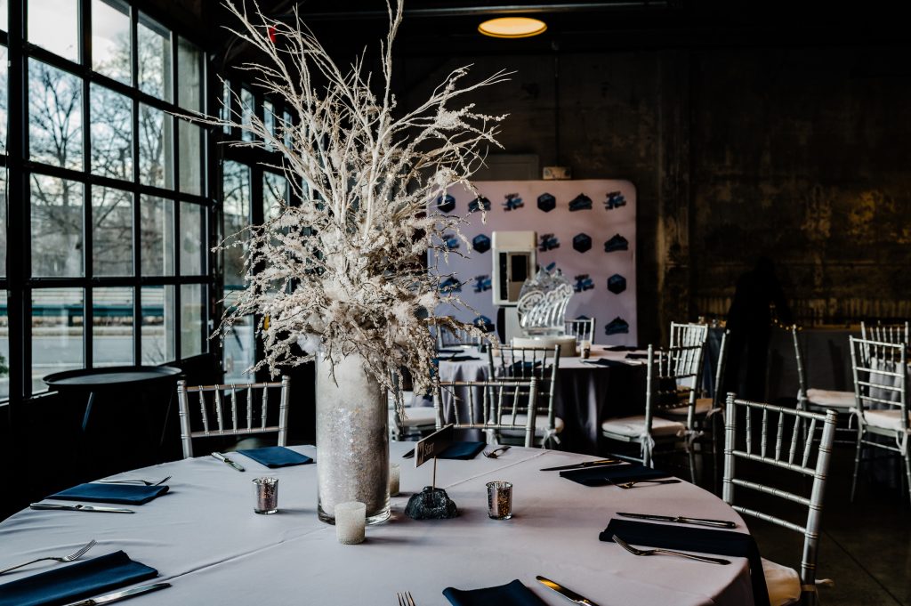 A round banquet table -- surrounded by silver Chiavari chairs -- topped with a large centerpiece comprised of flocked twigs in a glass container set up beside a floor-to-ceiling glass window against a backdrop of other tablescapes and a branded photo booth.