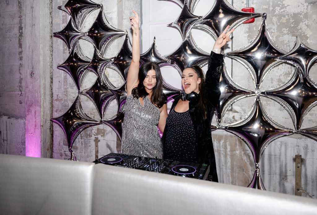 Two people dancing behind a DJ booth backed by star-shaped balloons against a distressed stone wall.