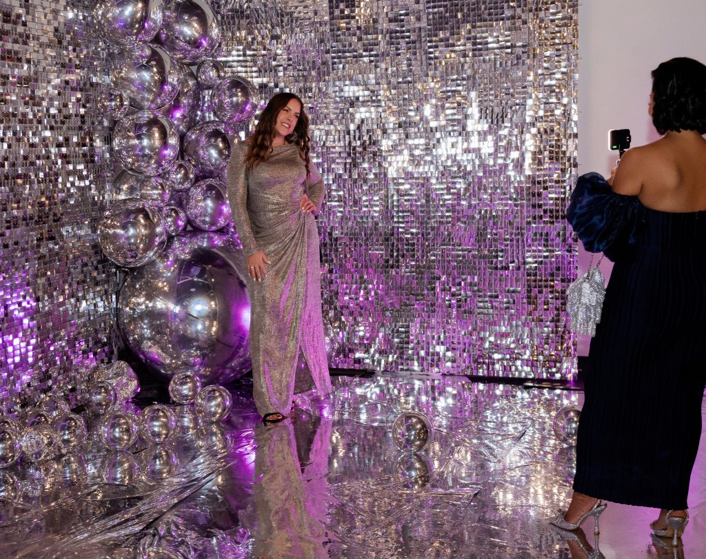 A person takes a photo of a woman in a floor-length silver gown against a glittering backdrop that features a chrome-colored balloon sculpture.
