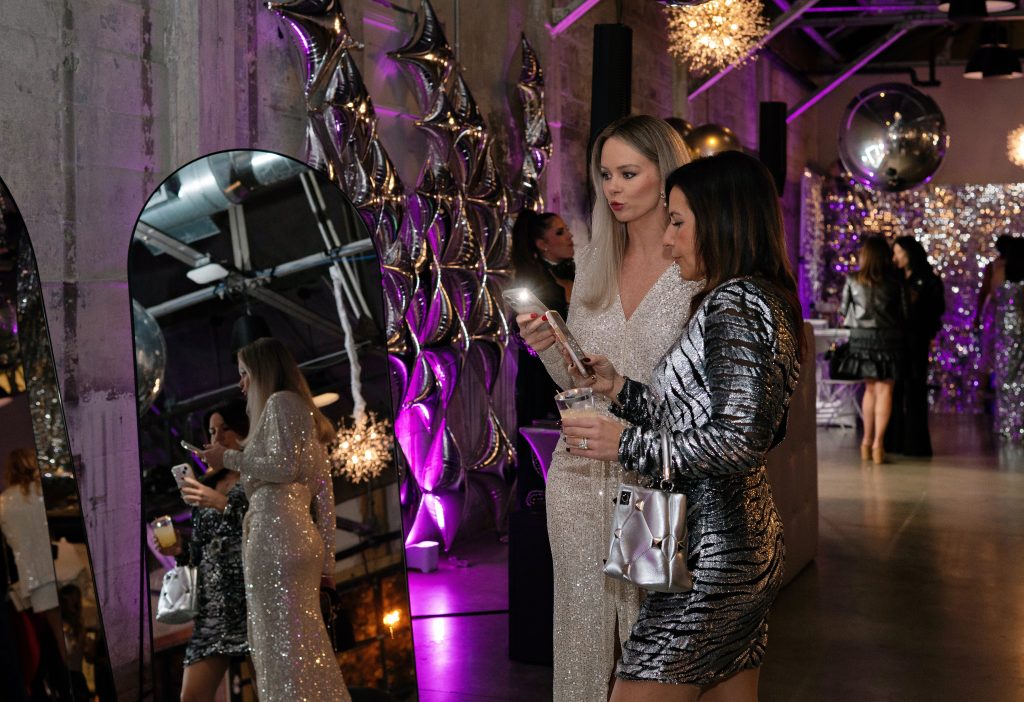 Two people dressed in silver evening attire take photos in front of a mirror at a wintry party held in a modern-industrial venue decorated with chrome accents.