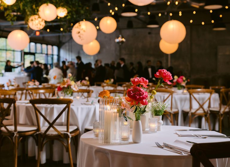 A modern-industrial venue outfitted for a wedding reception with paper lanterns, string lights, and greenery hanging from steel rafters at the ceiling as well as round banquet tables dressed with white linens and ikebana arrangements and arranged with wooden Chiavari chairs with a crowd of guests at a bar set against a large glass window in the background.