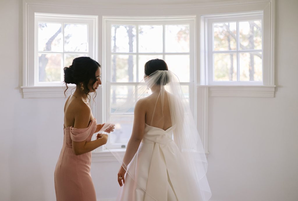 A bridesmaid assists a bride with her veil in front of a cottage-style window in a room with white walls.
