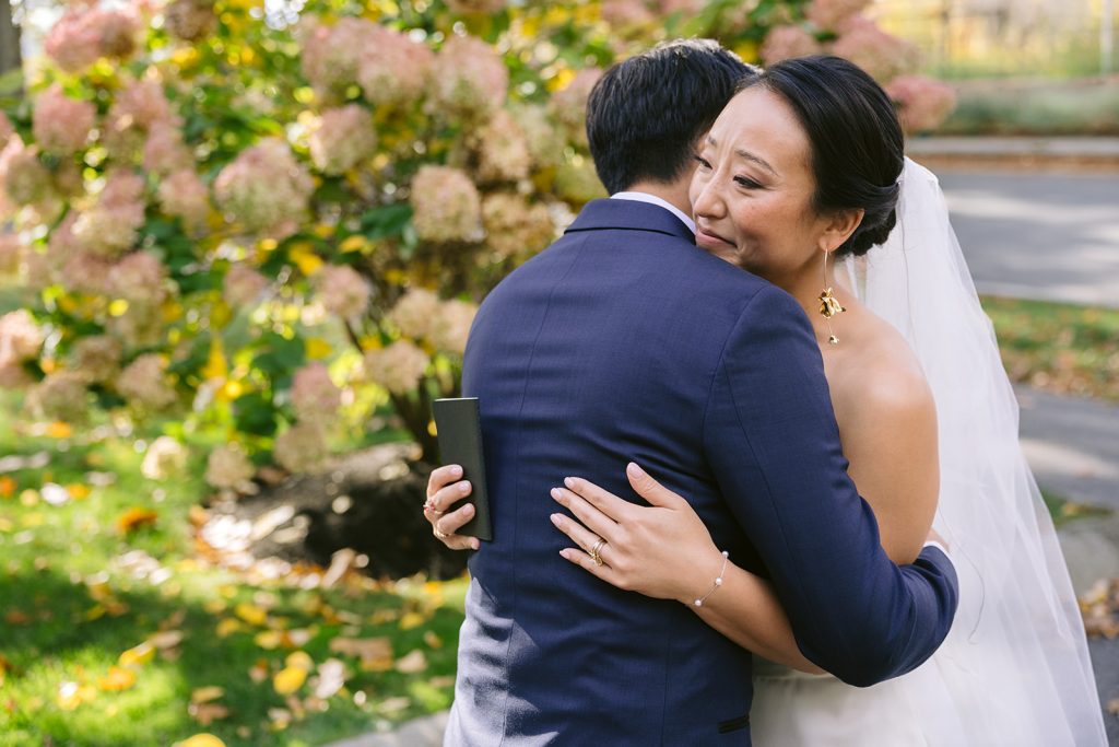 A groom hugs a happily tearful bride against a flowered hydrangea bush.