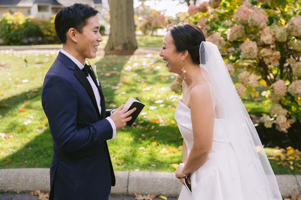 A smiling groom reads private vows to his laughing bride against a lawn landscape featuring a flowered hydrangea bush.