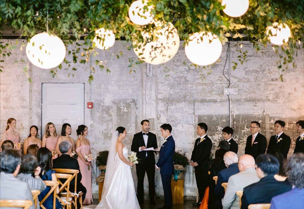 A wedding ceremony -- complete with bride and groom, officiant, and ten party members -- taking place against a distressed stone wall and underneath a ceiling arrangement of paper lanterns and greenery in front of rows of guests seated in wooden Chiavari chairs.