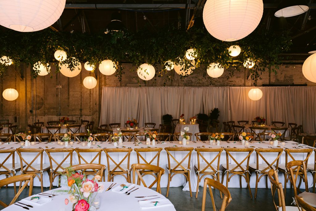A modern-industrial venue outfitted for a wedding reception with paper lanterns and greenery hanging from steel rafters at the ceiling as well as a layout of a long, rectangular table and a few round banquet tables dressed with white linens and ikebana arrangements and arranged with wooden Chiavari chairs against a pipe-and-drape installation lining a distressed stone wall.