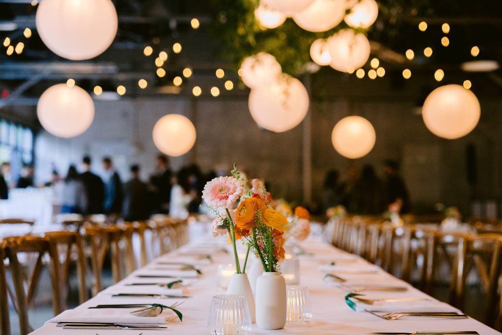 A modern-industrial venue outfitted for a wedding reception with paper lanterns, string lights, and greenery hanging from steel rafters at the ceiling, featuring a long, rectangular table dressed with a white linen and ikebana arrangement and arranged with wooden Chiavari chairs in the foreground and mingling guests in the background.