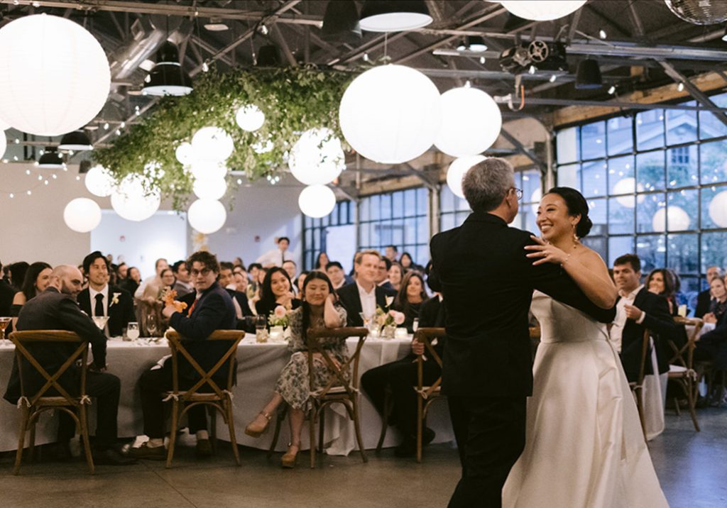 A bride dances with her father in front of wedding guests seated at tables underneath a ceiling arrangement of paper lanterns and greenery hanging from steel rafters in a modern-industrial venue that features a bank of floor-to-ceiling glass windows.