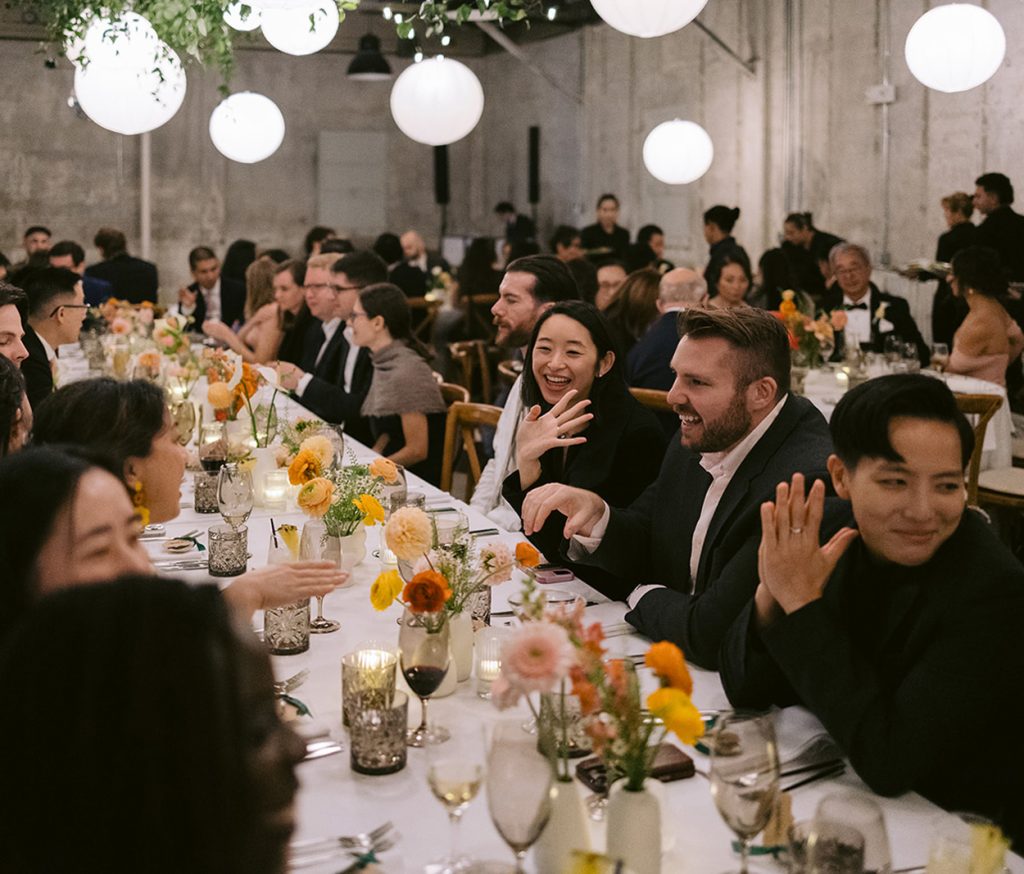 Smiling people talk to each other along a rectangular table at a wedding reception taking place in a modern-industrial venue featuring distressed stone walls and hanging paper lanterns.