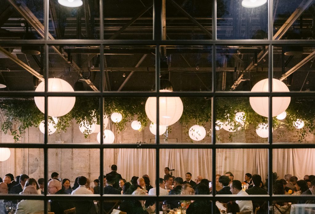 A view through a large glass window and its muntins into a modern-industrial venue filled with wedding guests underneath a ceiling arrangement of paper lanterns and greenery that also features a pipe-and-drape installation in front a distressed stone wall.