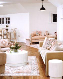 Neutral- and pastel-toned furniture -- including a high-top table and chairs, sofas, and accent tables -- arranged in a room with white walls and natural hardwood flooring featuring a rounded turret corner at one wall.